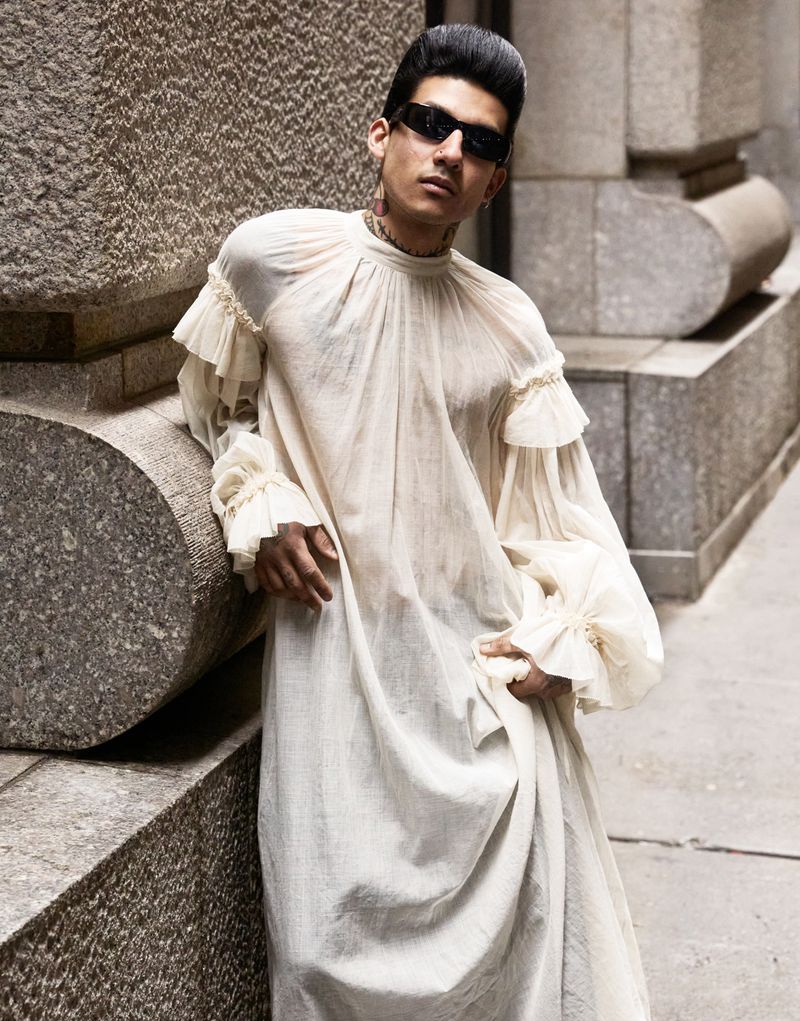 A stylish adult male poses in a long white dress with ruffles, sunglasses, and a high pompadour hairstyle outdoors in an urban setting.