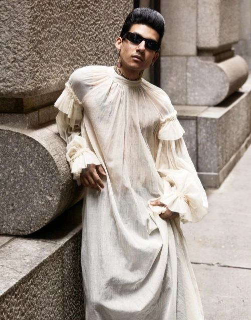 A stylish adult male poses in a long white dress with ruffles, sunglasses, and a high pompadour hairstyle outdoors in an urban setting.