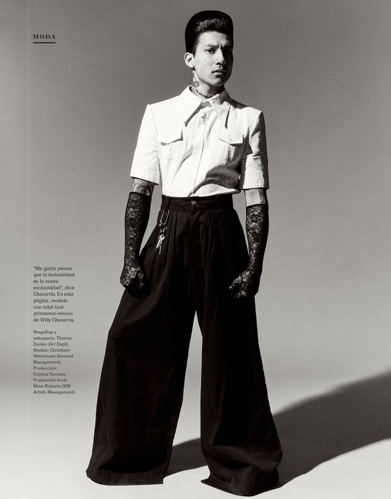 Editorial fashion shot of a slim male model with tattoos posing in a white short-sleeved shirt and black high-waisted wide-legged pants.