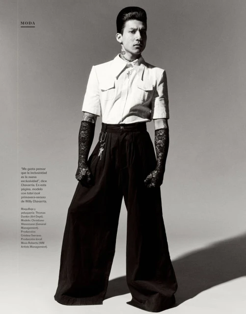 Editorial fashion shot of a slim male model with tattoos posing in a white short-sleeved shirt and black high-waisted wide-legged pants.