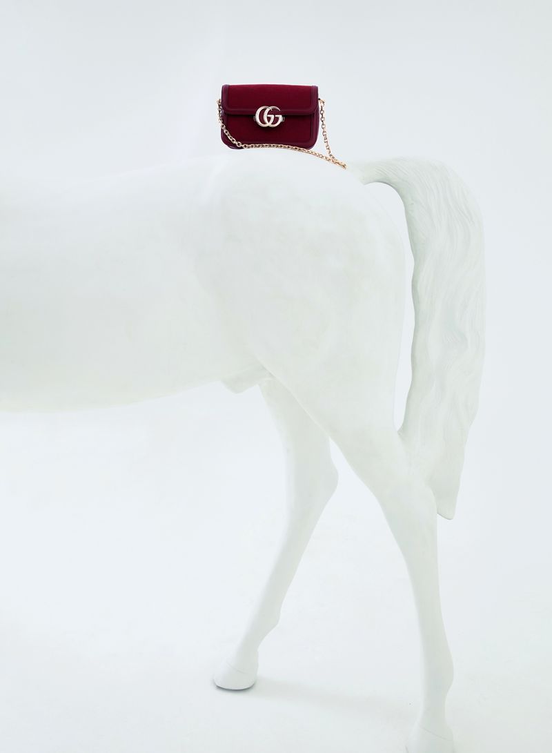 Burgundy Gucci purse with a gold chain strap rests on the back of a matte white horse sculpture against a white background.