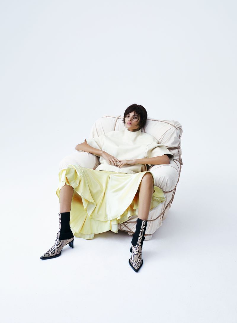 A woman in a cream top and yellow skirt sits in a modern chair against a white backdrop, showcasing a relaxed fashion aesthetic.