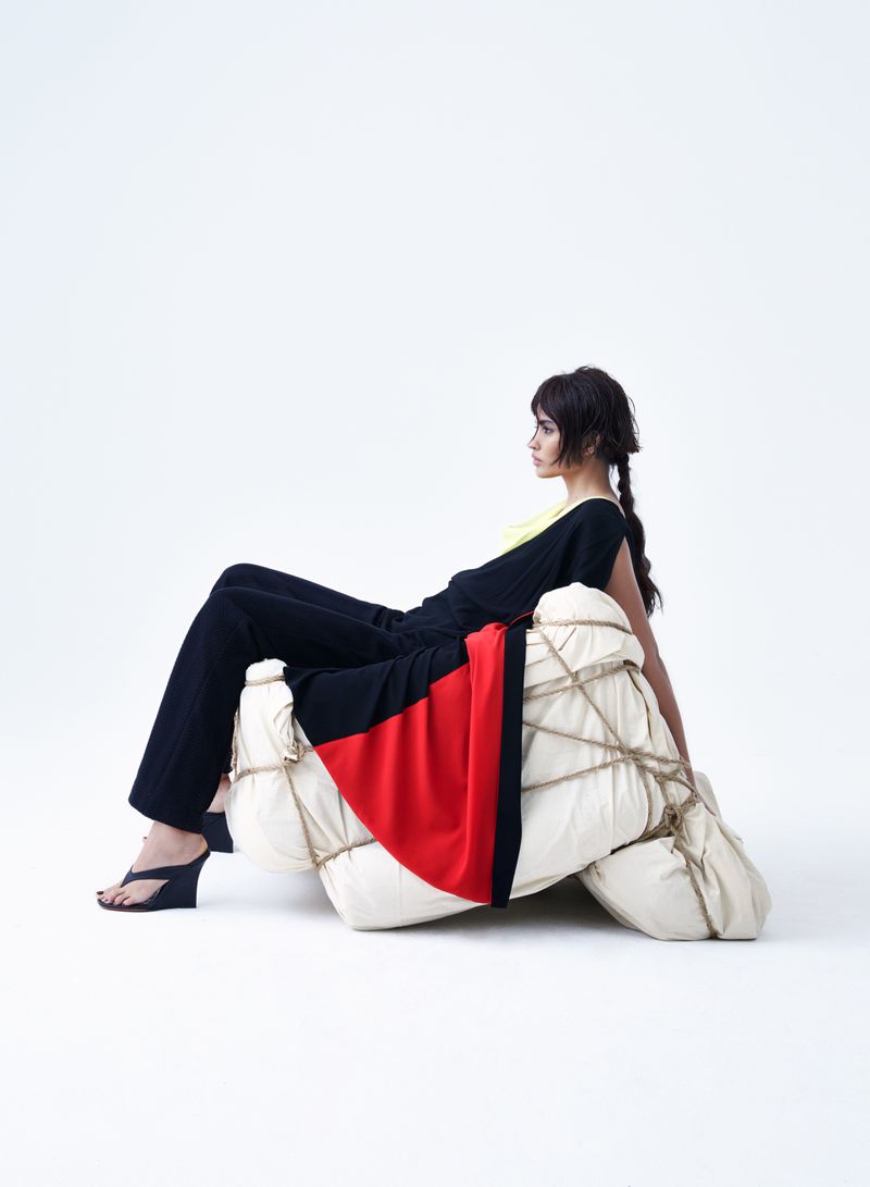 Woman seated on wrapped objects in a studio, wearing a black top and pants with high heels. The background is bright white.