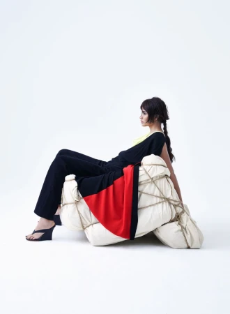 Woman seated on wrapped objects in a studio, wearing a black top and pants with high heels. The background is bright white.