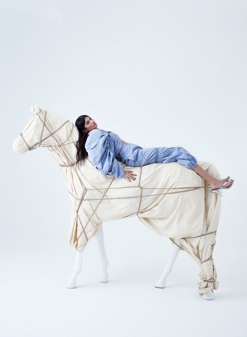 A brunette woman wearing a light blue outfit is reclining on a toy horse wrapped in string on a white seamless background.