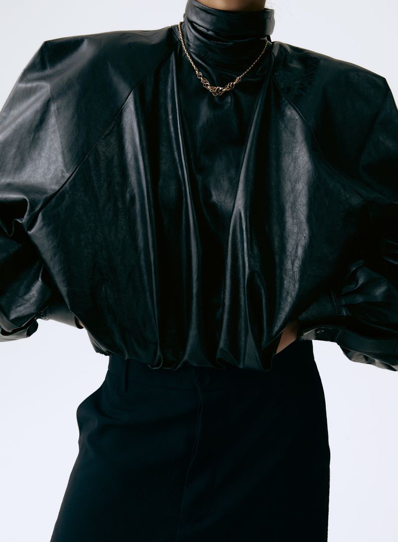 Model wears a black leather top with puffy sleeves and black pants against a white backdrop, showcasing fashion and style.