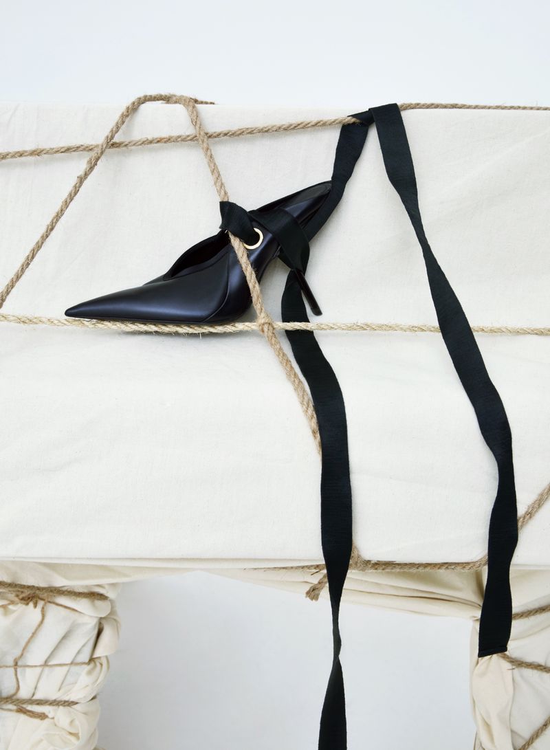 Elegant black high heel pump with a pointed toe and ribbon accents, displayed on a fabric surface tied with jute rope and black ribbon.