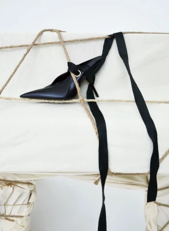 Elegant black high heel pump with a pointed toe and ribbon accents, displayed on a fabric surface tied with jute rope and black ribbon.