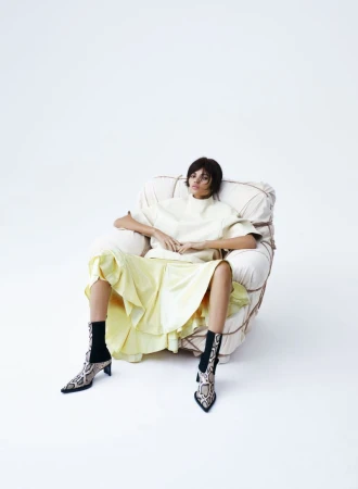 Model seated in an armchair wearing a light top, yellow skirt, and snakeskin boots against a white background
