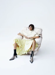 Model seated in an armchair wearing a light top, yellow skirt, and snakeskin boots against a white background
