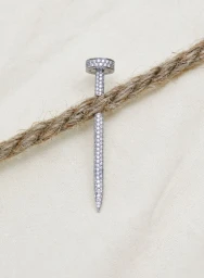 Diamond encrusted nail shaped jewelry piece resting over a thick braided rustic rope against a neutral textured background.