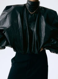 Model wearing a black leather high-neck top with structured shoulders and a gold chain necklace against a white background.