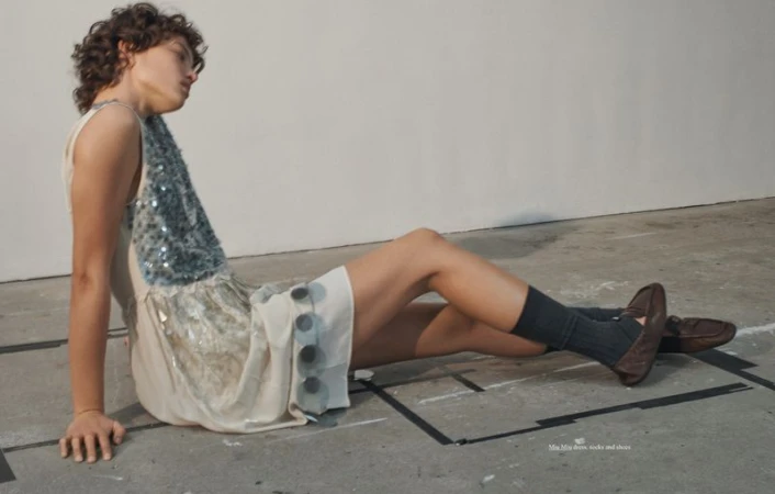 Model seated on concrete floor wearing a sleeveless dress with sequin panel, dark socks, and brown loafers