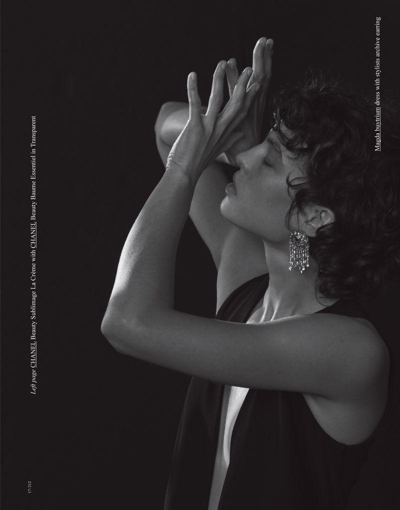 Black and white studio shot of a young adult woman in a dress, raising her hands with chandelier earrings against a dark background.