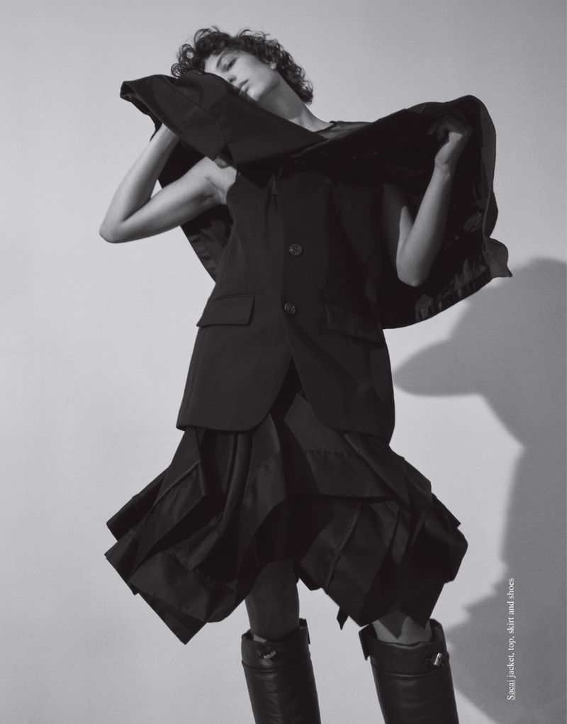 Model with short curly hair posing in a black blazer and flared skirt against a light studio backdrop for Love Want Magazine.