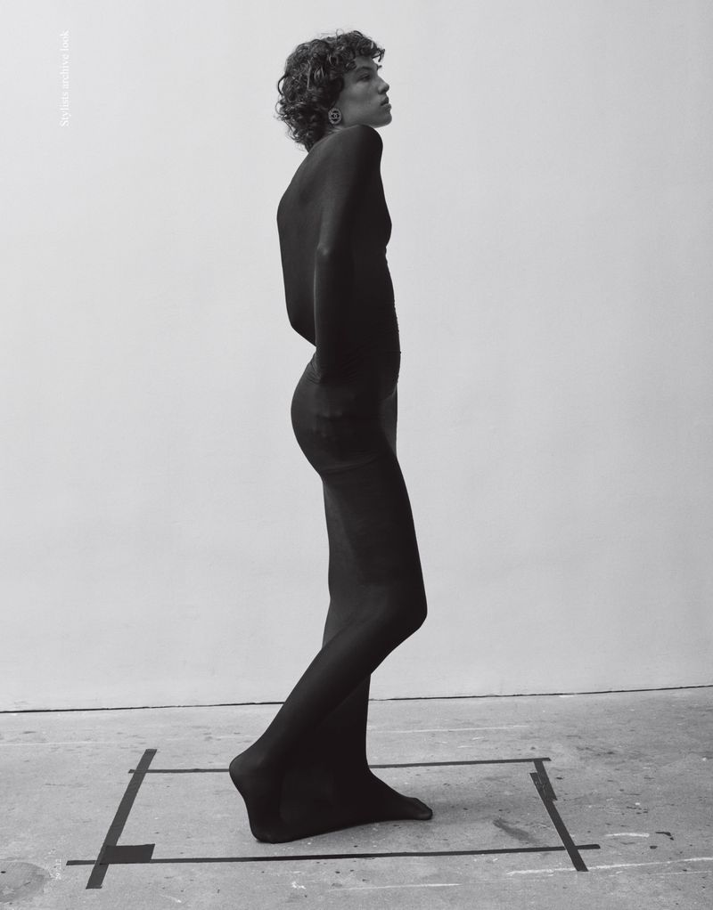 A curly-haired person in a black dress walks across a grey floor marked with black tape, set against a plain white backdrop.