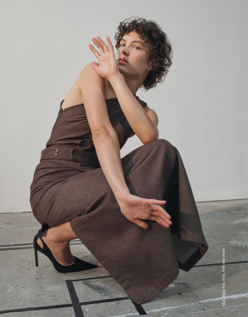 A slender woman with curly hair poses in a one-shoulder brown jumpsuit and black heels in a studio with Mondrian-inspired floor art.