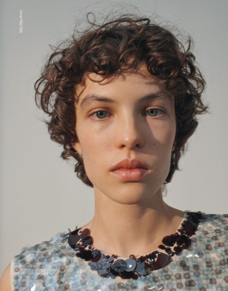 Close-up portrait of a fair-skinned adult female with curly brown hair and blue eyes, wearing a sequined dress with dark trim on white backdrop.