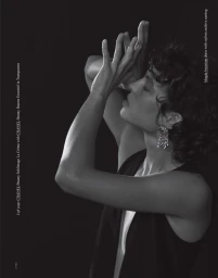 Black and white editorial portrait of a woman posing with hands raised near her face against a dark background.