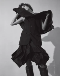 Model with short curly hair posing in a black blazer and flared skirt against a light studio backdrop for Love Want Magazine.