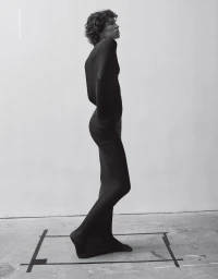 Model with short curly hair wearing full black body tights standing in profile inside taped floor markings