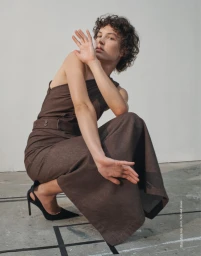 Model with curly hair squatting in a brown belted top and skirt with black heels against a neutral background