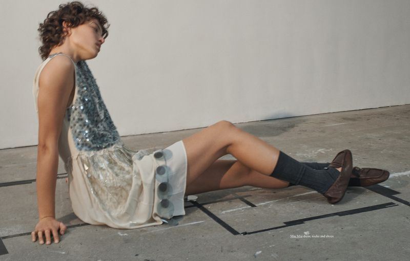 Model seated on concrete floor wearing a sleeveless dress with sequin panel, dark socks, and brown loafers