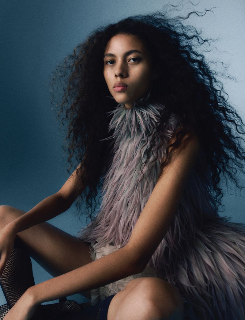 Portrait of a woman with dark, curly hair wearing a feathered garment against a blue background in a beauty and fashion editorial style.