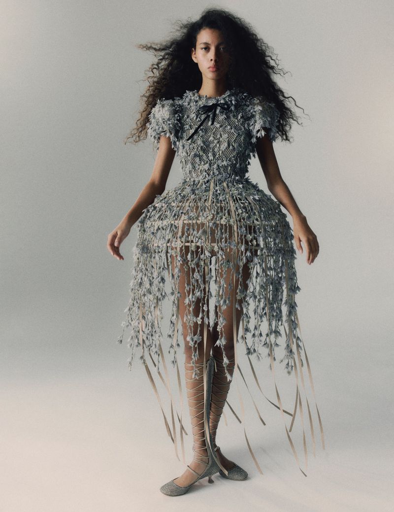 A slender young woman with curly dark hair poses in a couture dress made of shimmering grey material with matching Mary Jane heels.