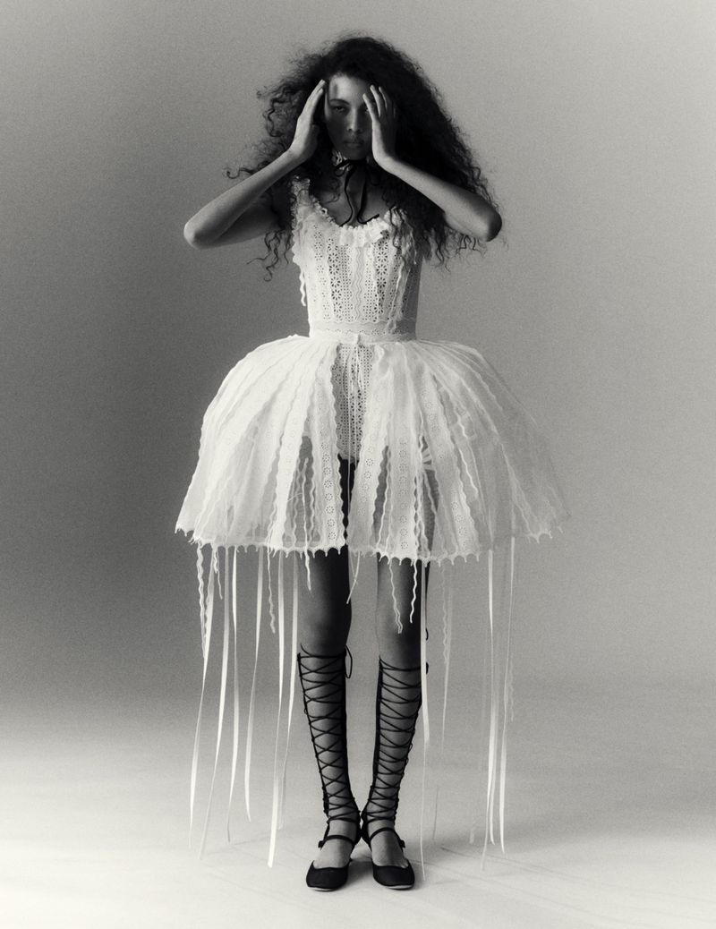 Monochrome portrait of a woman in a lacy dress and tall sandals, standing against a studio backdrop, her hands on her head.