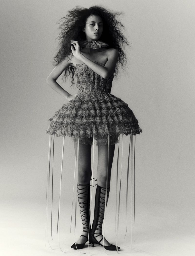 Full-length portrait of a woman wearing a dress with ribbons, gladiator-style shoes, and curly hair, set against a neutral background.