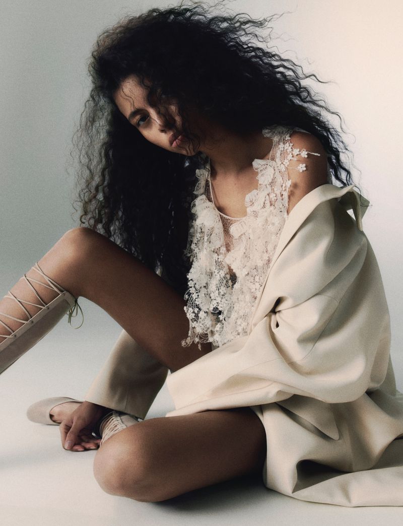 Adult woman with curly black hair sits indoors wearing a beige jacket and lace top, posing against a neutral background in a studio shot.