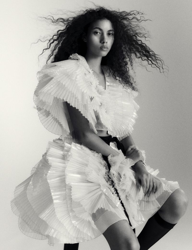Black and white portrait of a woman wearing a ruffled dress and knee-high socks in a photo studio setting with soft lighting.