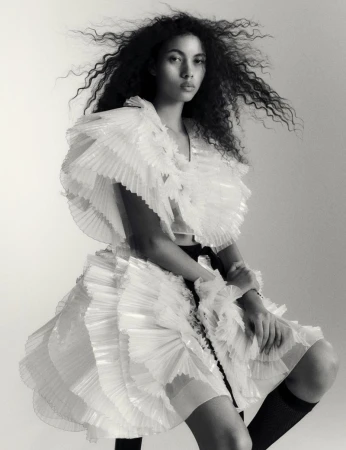 Black and white portrait of a woman wearing a ruffled dress and knee-high socks in a photo studio setting with soft lighting.