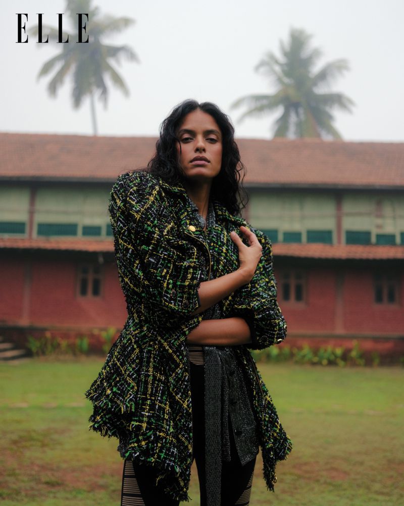 A woman in a patterned jacket poses outdoors with a red-roofed building and palm trees in the soft background, editorial fashion photography.