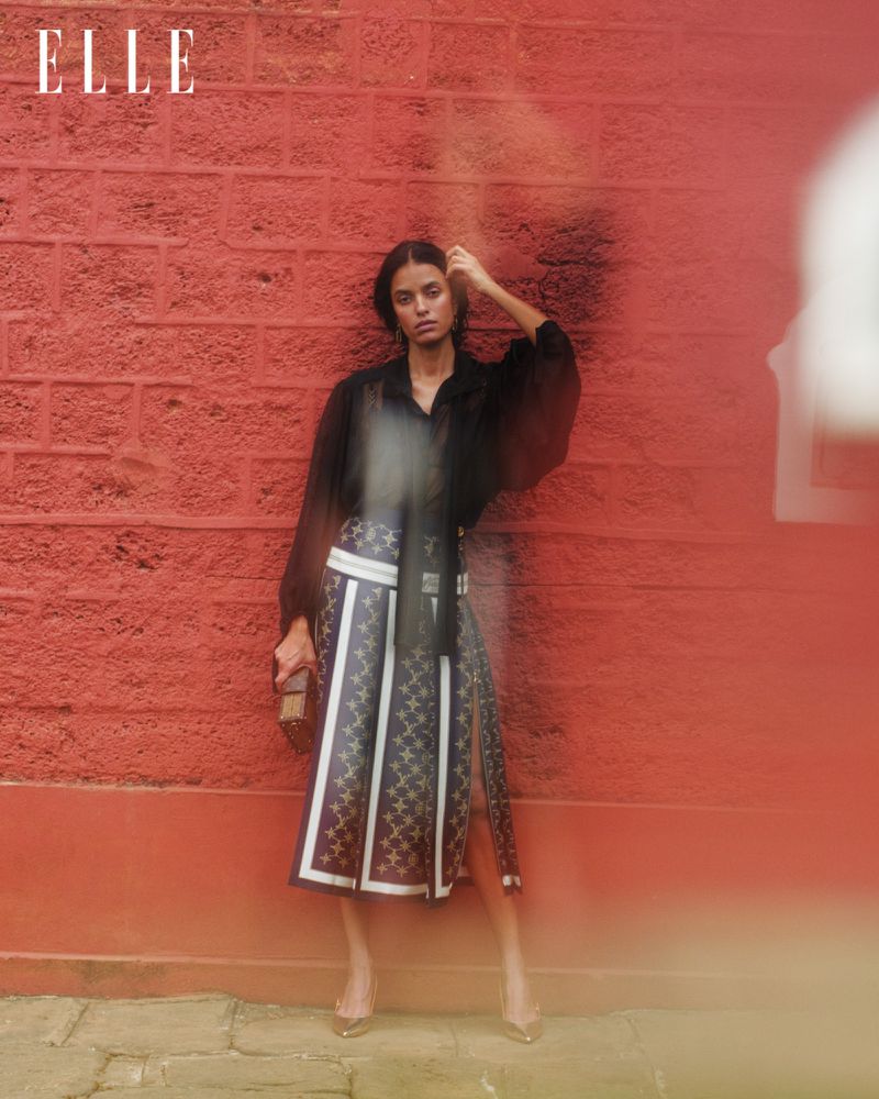 A stylish adult female model stands in front of a brick wall, wearing a black blouse, patterned skirt, and high heels, holding a clutch.