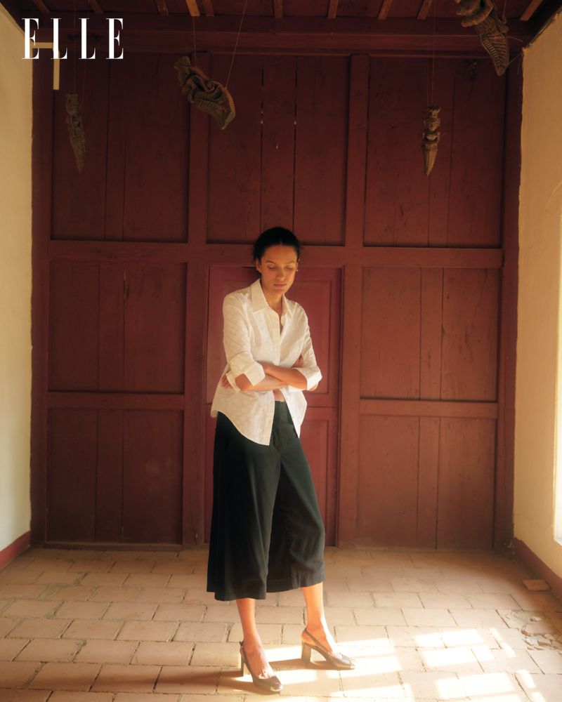 Elegant woman in a white shirt and black culottes standing in a hallway with a tiled floor and dark wood paneling.