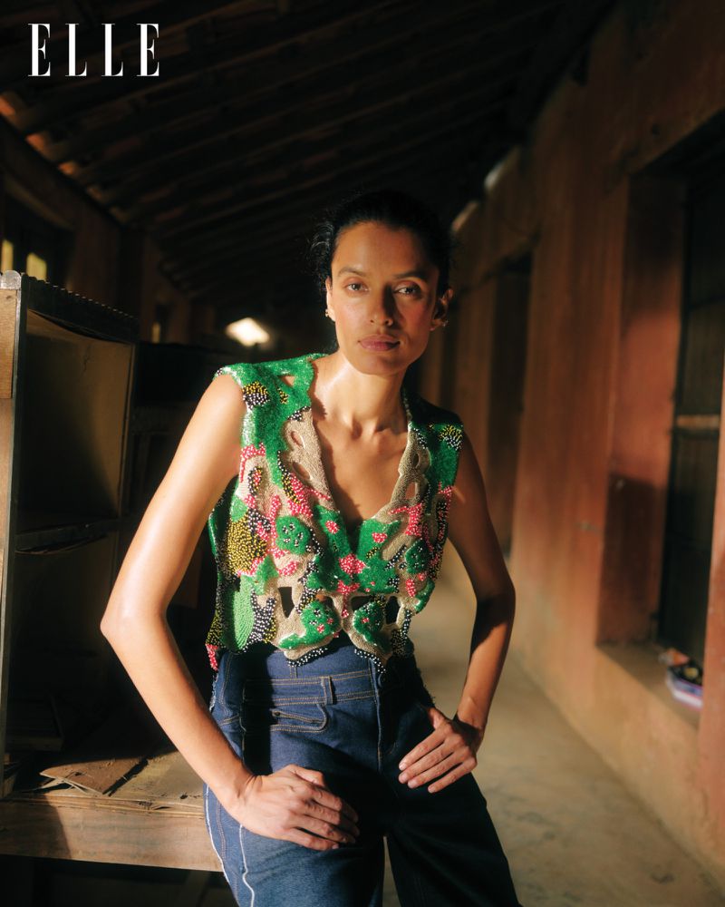 Adult female poses indoors wearing a green beaded top and blue jeans, standing with hands on hips in a medium shot with soft lighting.