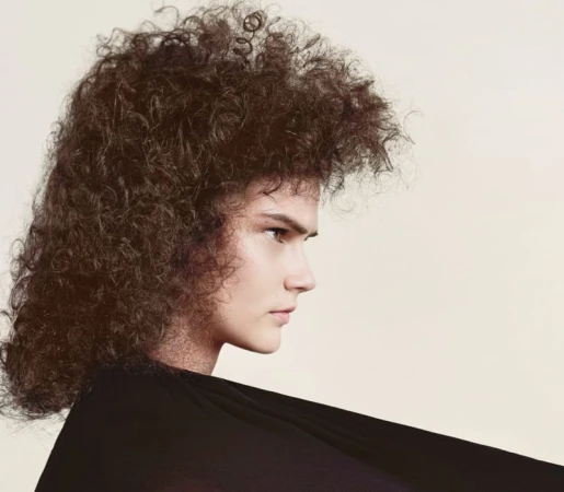 Profile shot of a woman with curly brown hair, wearing a black top, set against a light backdrop in a beauty or fashion context.