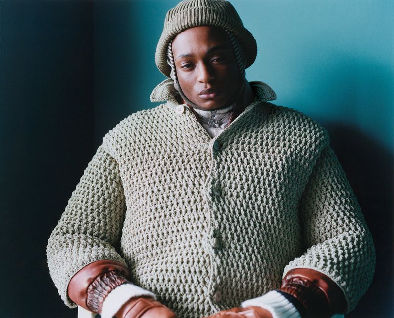 Fashionable African-American man in a knitted hat and open knit cardigan with leather gloves poses indoors against a teal-colored wall.