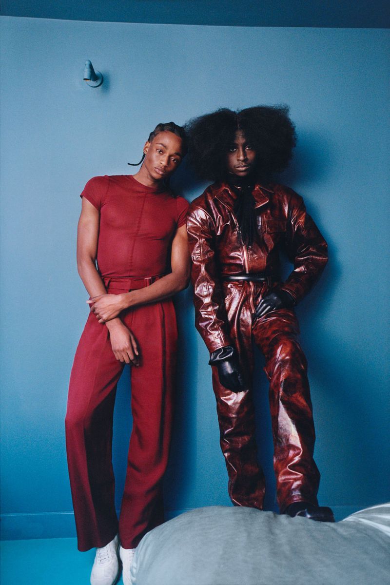 Two figures in bold red and brown leather outfits pose indoors against a muted blue wall on a puffy grey surface, fashion-forward and confident.