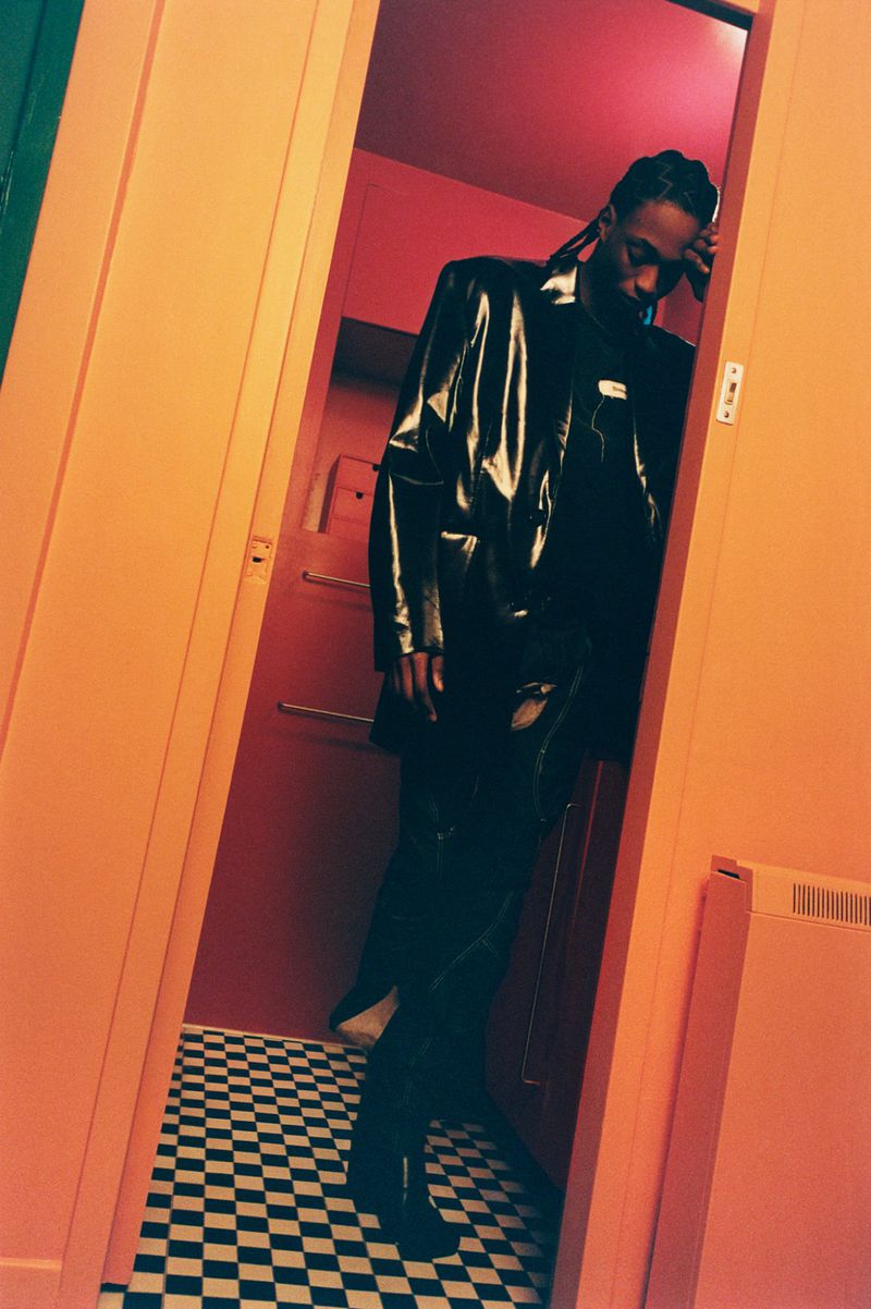 A Black male in a shiny coat leans in an orange doorway with a red wall and black and white checkered floor in the background.