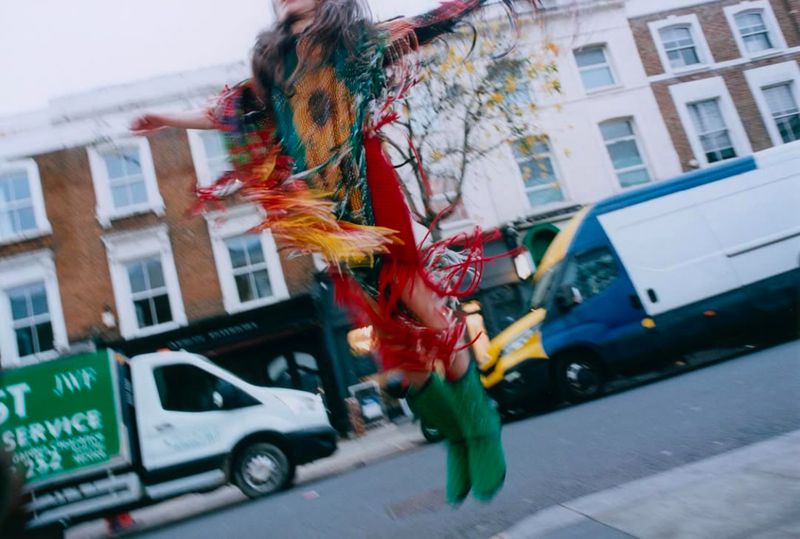A young woman in a colorful fringed outfit mid-air on a city street, with buildings and trucks visible in the background, motion blur present.