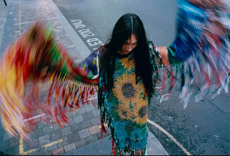 Person with long hair and vibrant wings stands on a city street, wearing a sunflower-patterned top and a unique garment with fringes.