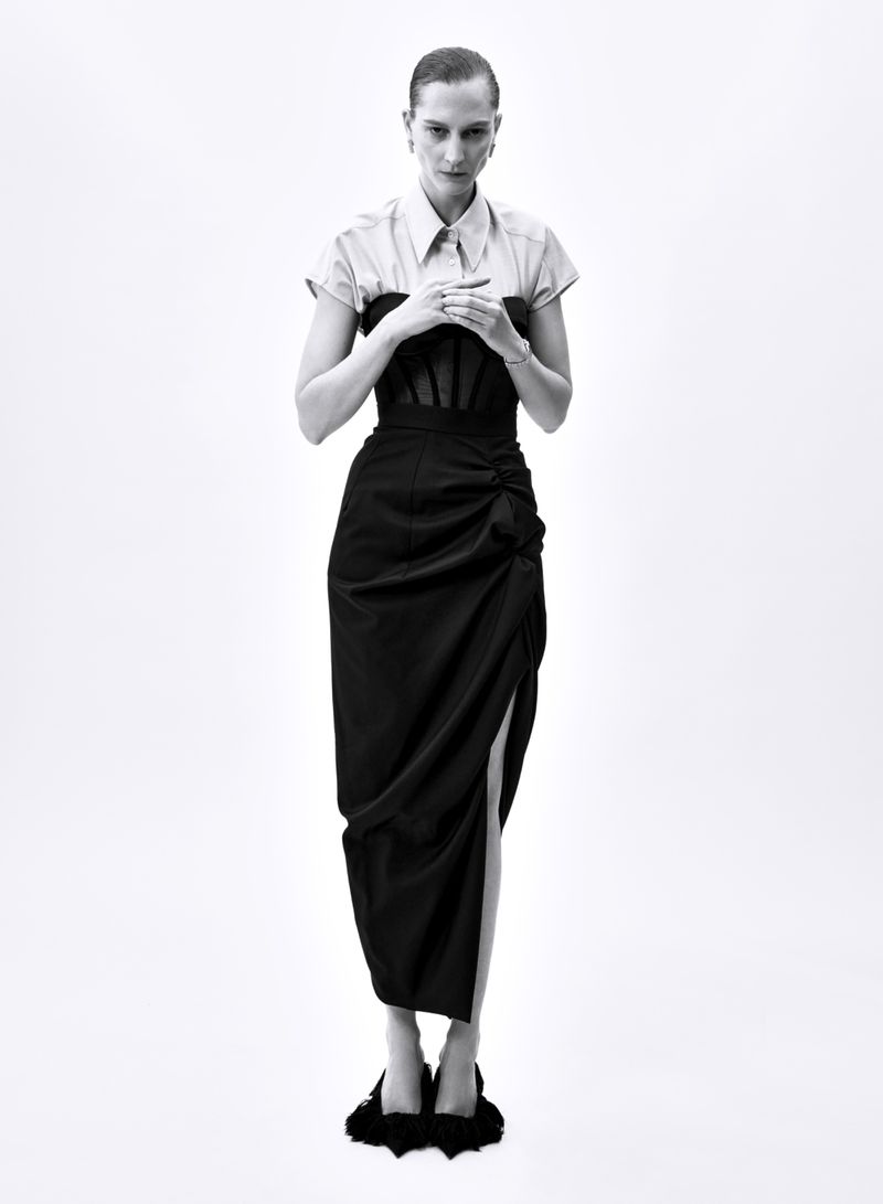 Black and white studio shot of a woman in a collared shirt, black corset, and long skirt with slit, posing against a white backdrop.