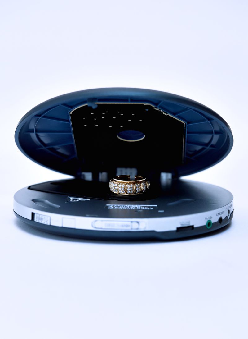 Elevated shot of a gold ring with gemstones resting inside an open portable compact disc player against a bright white background.