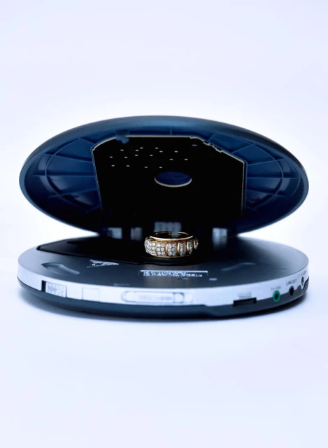 Elevated shot of a gold ring with gemstones resting inside an open portable compact disc player against a bright white background.