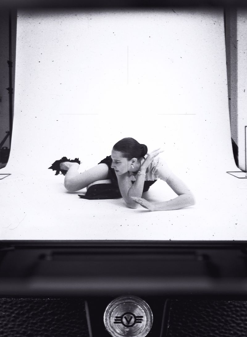 A woman poses on a white background in a photo studio, captured through the viewfinder of a vintage camera, creating a retro aesthetic.