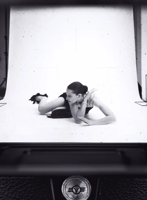 A woman poses on a white background in a photo studio, captured through the viewfinder of a vintage camera, creating a retro aesthetic.