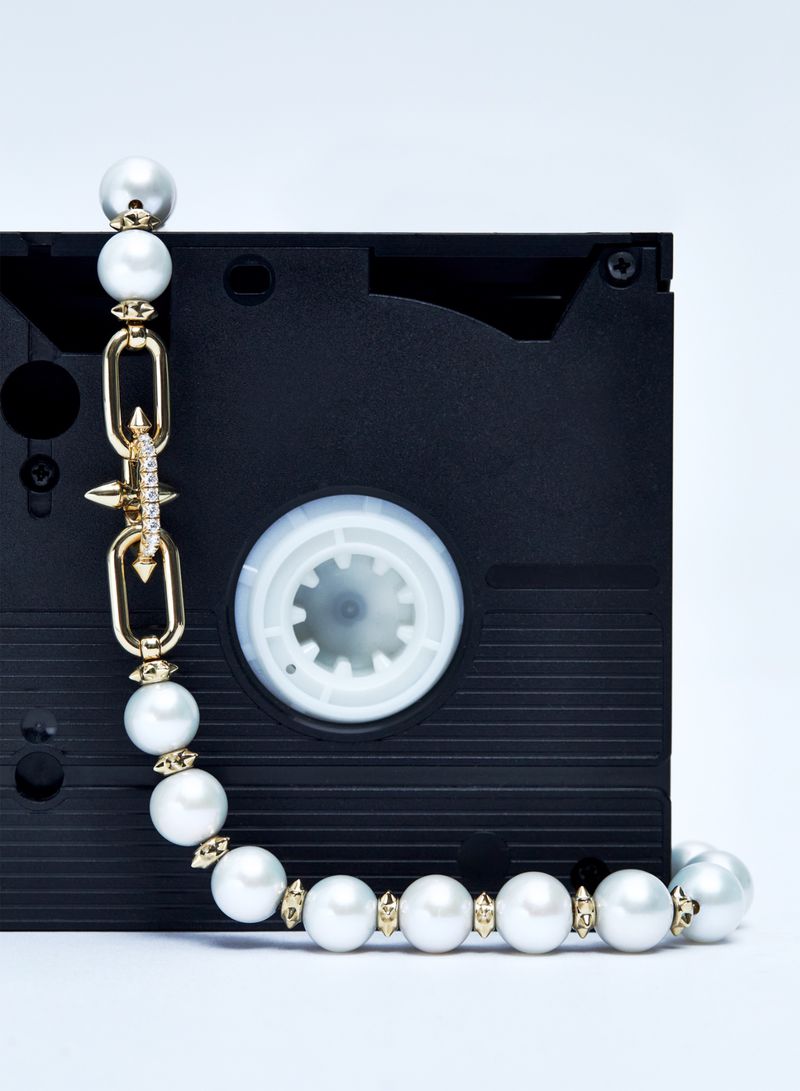 A pearl necklace with gold accents is draped over a black VHS tape against a white background in a studio setting, showcasing fashion jewelry.